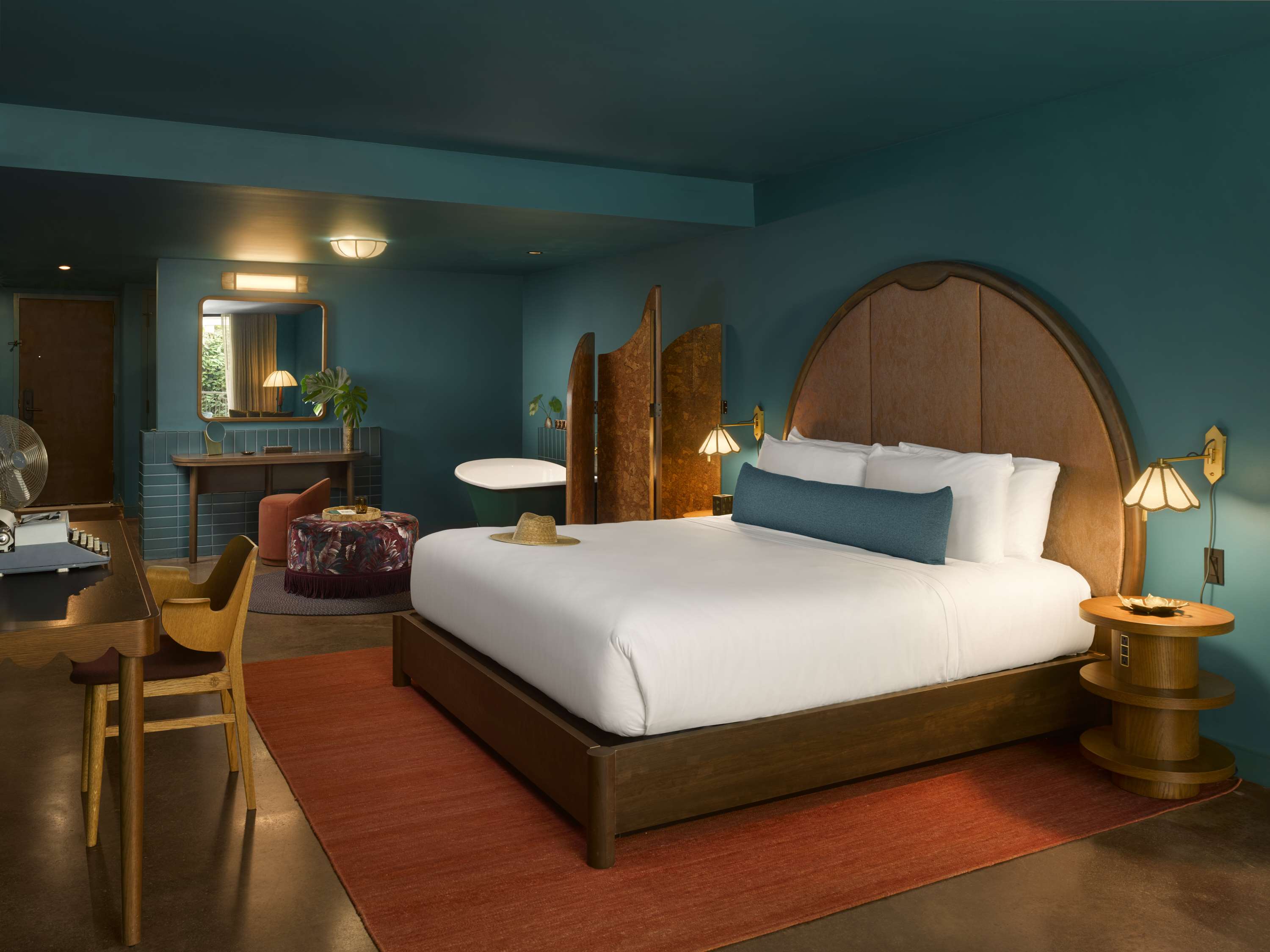 image King bed with a wood headboard and white linens, with a tropical pouf, wooden desk, and decorative screen in the background