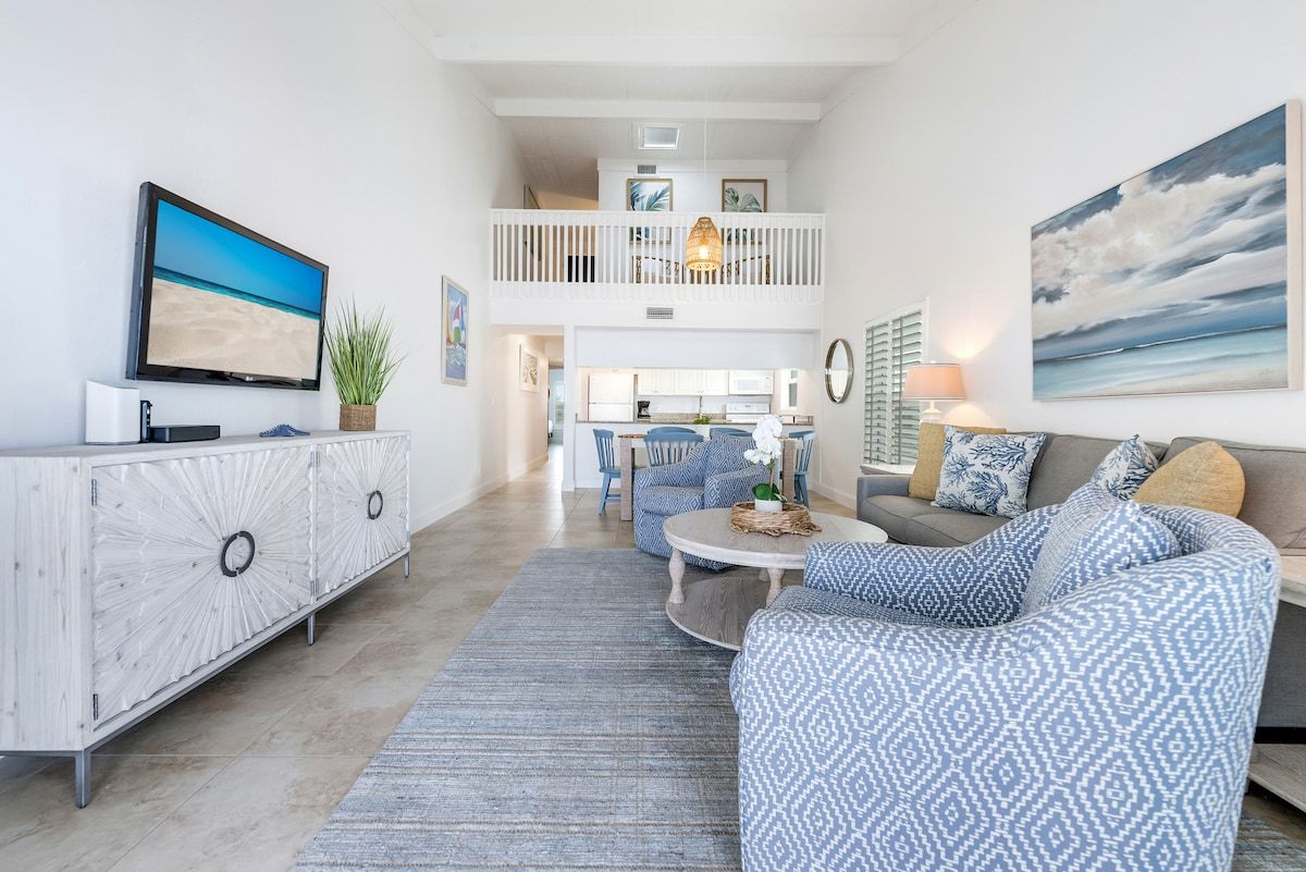 image Another view of the open-concept living area shows coastal décor, comfortable seating, a TV, and views of the kitchen and loft.