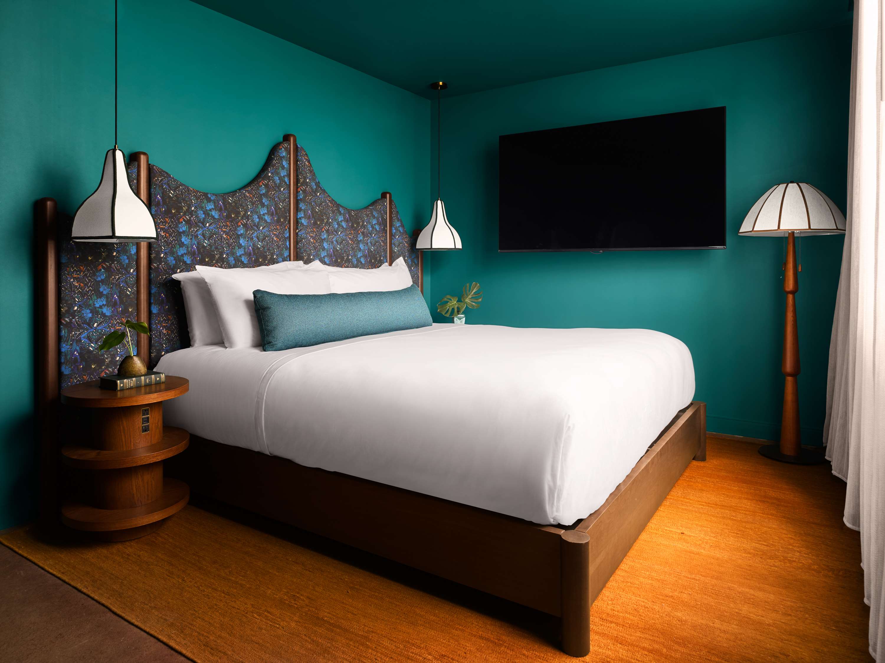image Teal-walled king bedroom with a patterned headboard, white duvet, fabric pendant lamps, and a wall-mounted flat-screen TV