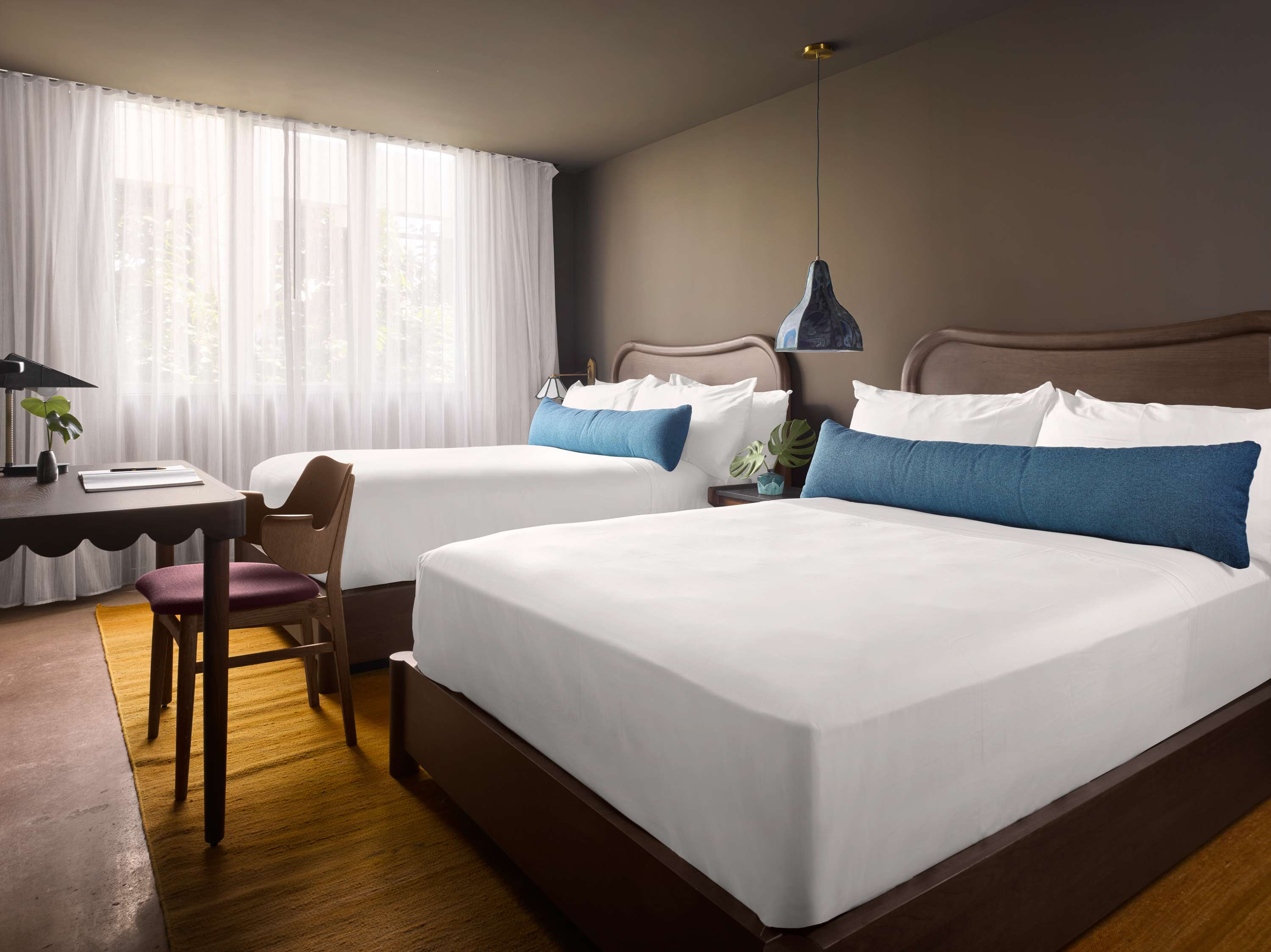 image Second bedroom with two beds, white linens, blue accent pillows, a wooden desk with a black lamp, and sheer curtains