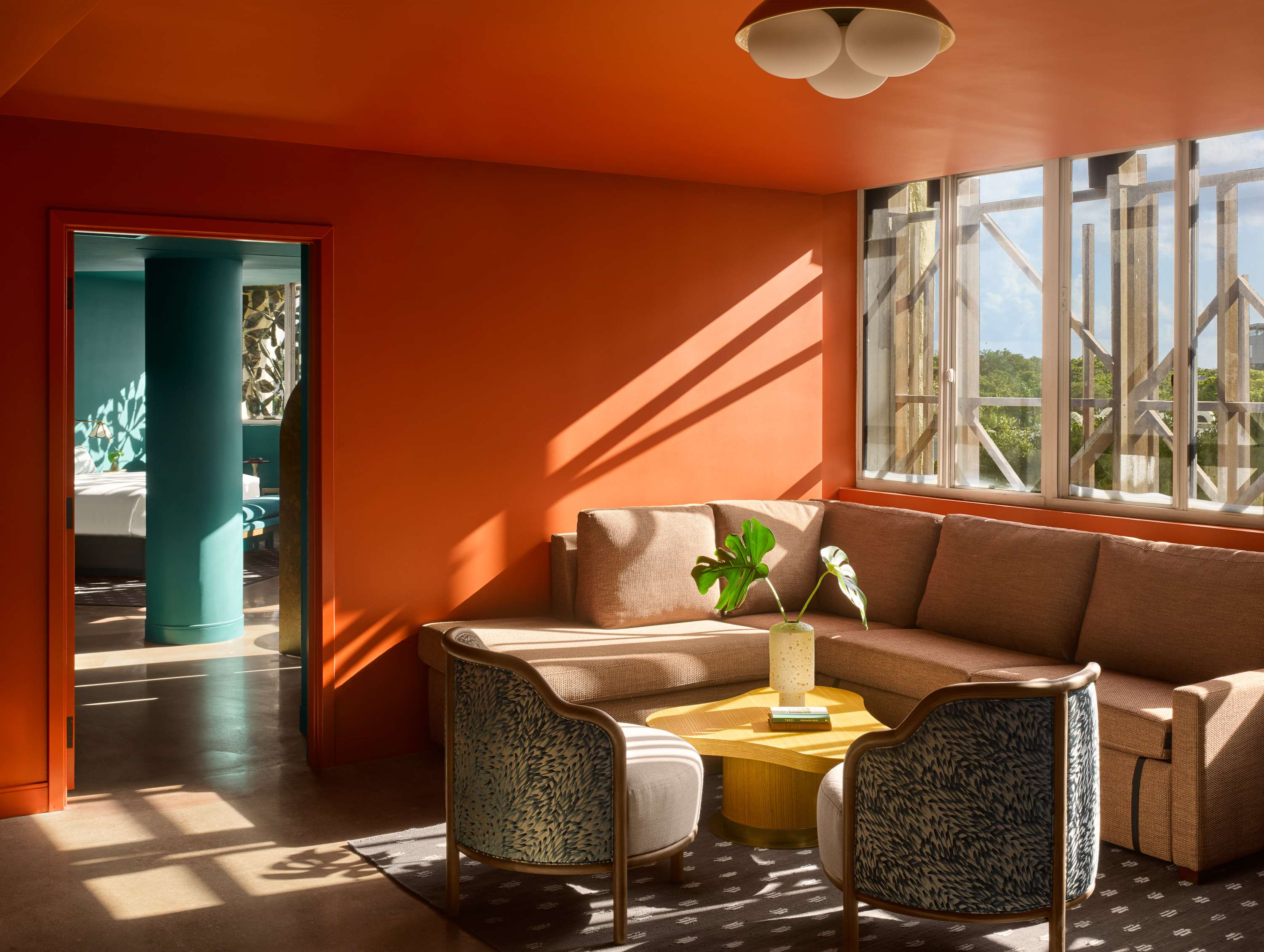image Sunlit living area with coral walls, patterned armchairs, and a yellow coffee table — the teal bedroom visible through the open doorway