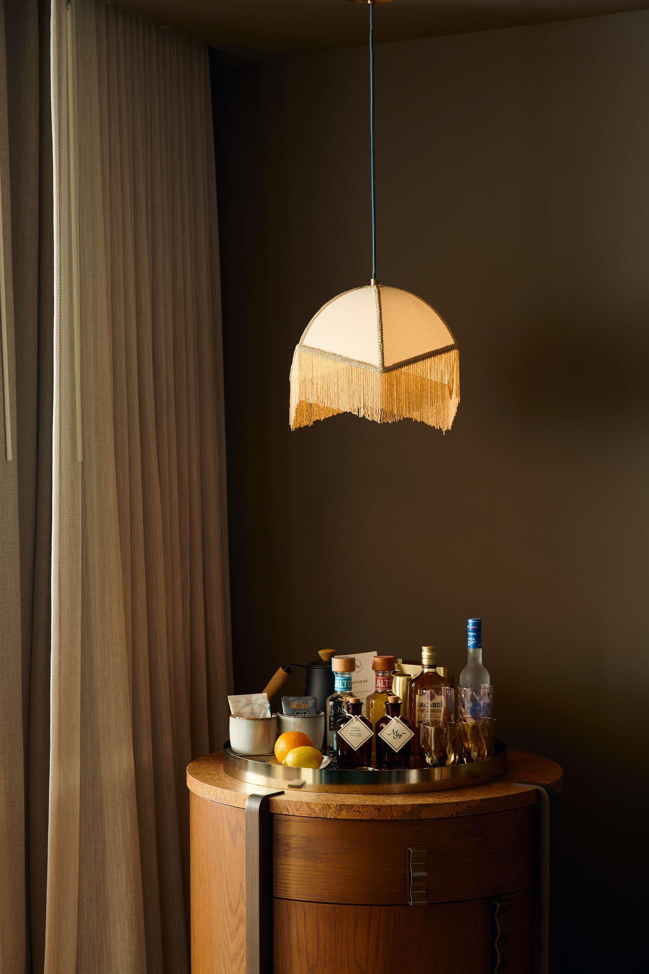 image Walnut bar cabinet stocked with spirits and mixers, topped by a gold fringe pendant lamp