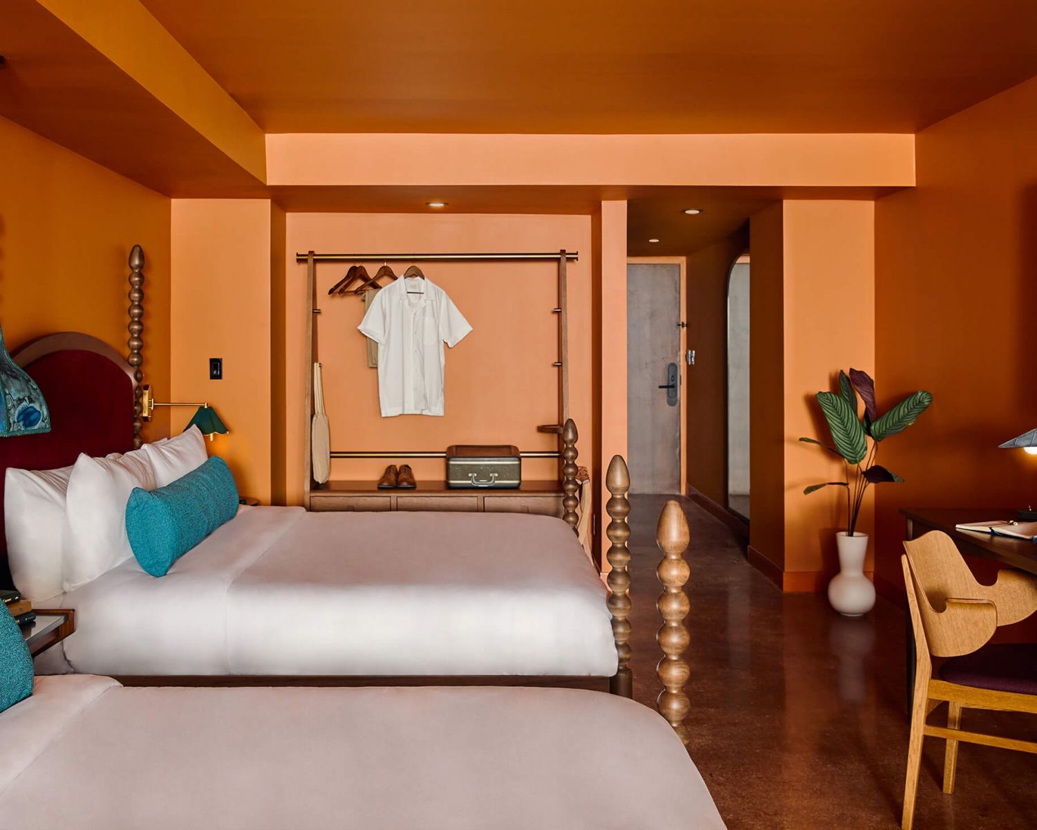 image Two beds with beige bedding and an aquamarine bolster, set against orange walls with a wooden dresser and hanging clothes rack