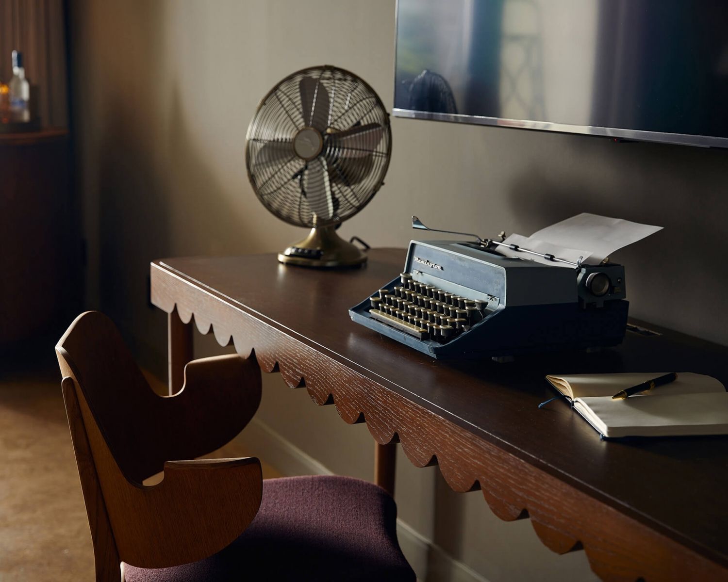 image Close-up of the scallop-edged mahogany desk with a vintage Remington typewriter, brass fan, and open notebook