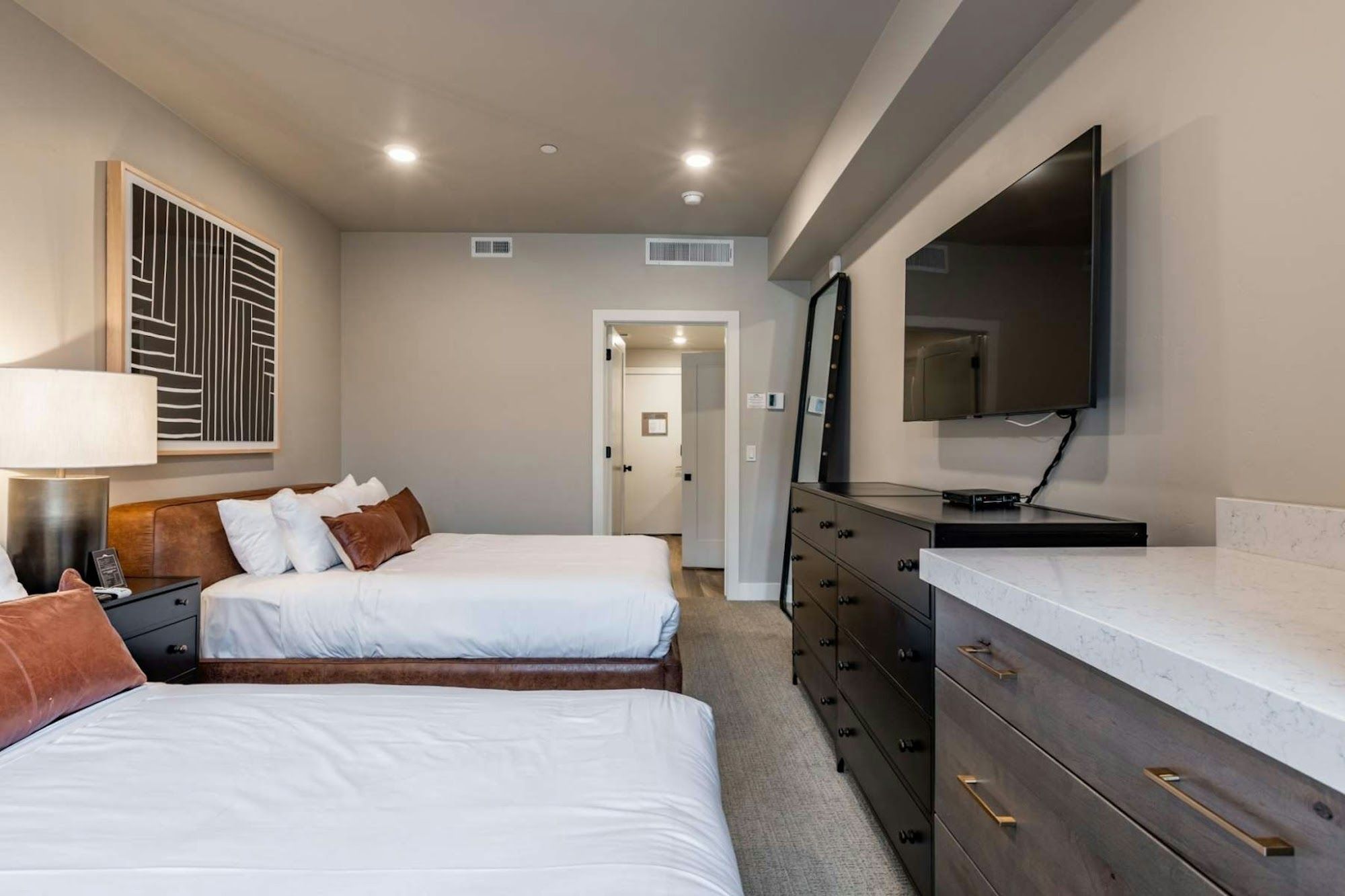 image Two queen beds with leather headboards, wall-mounted TV, dark dressers, and marble-top wet bar counter