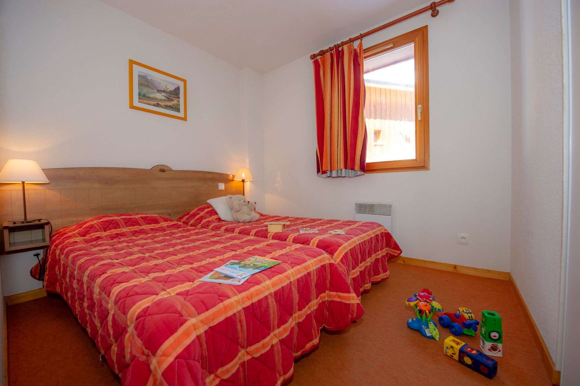 image A bedroom with two beds, a window, and children's toys on carpeted floor.