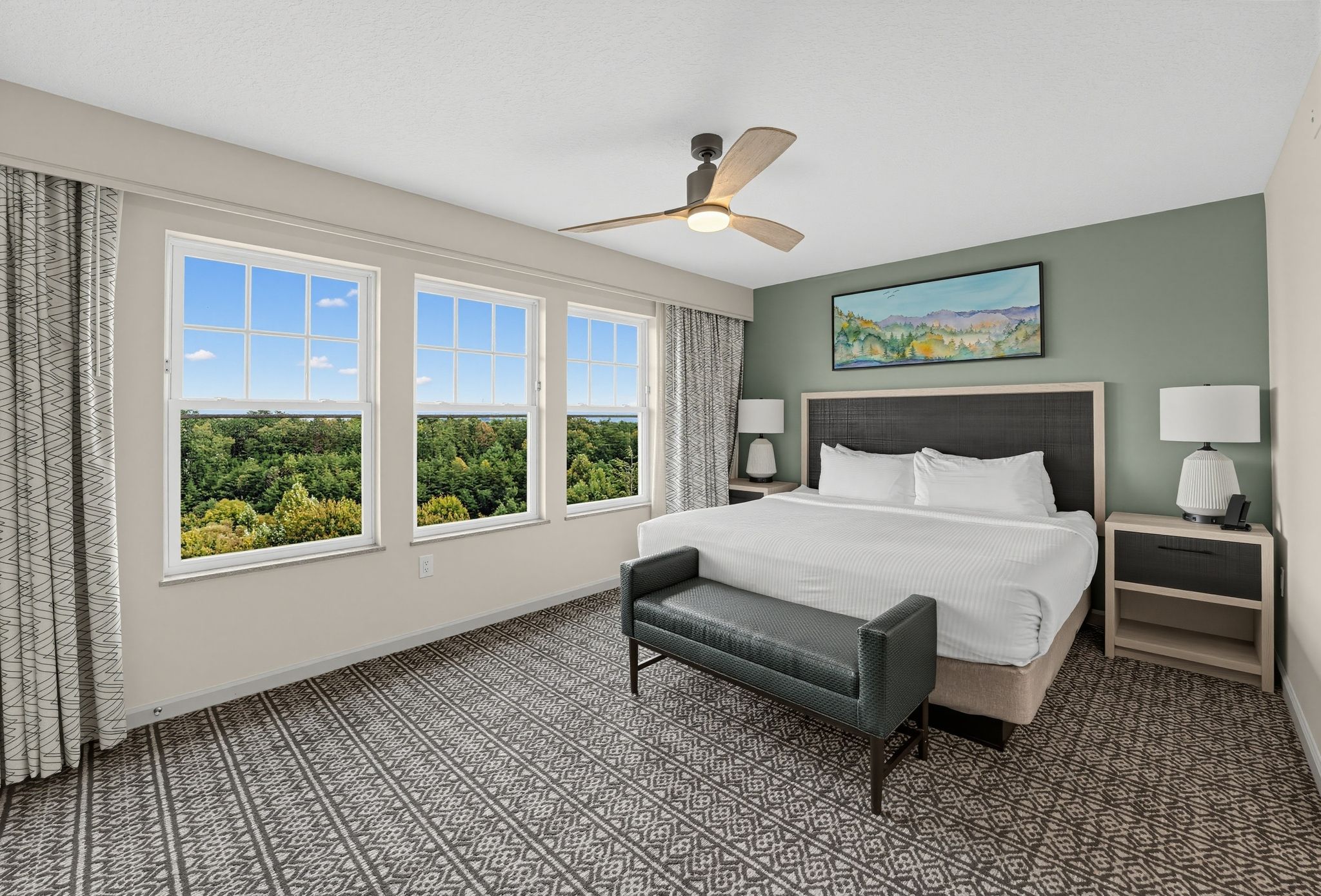 image Bedroom with a large bed and bench, nightstands with lamps, and windows overlooking trees.