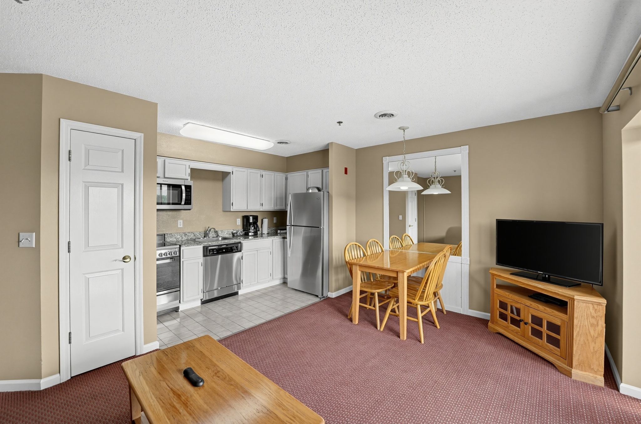 image Open living area with a TV, dining table with chairs, and a kitchen beyond.