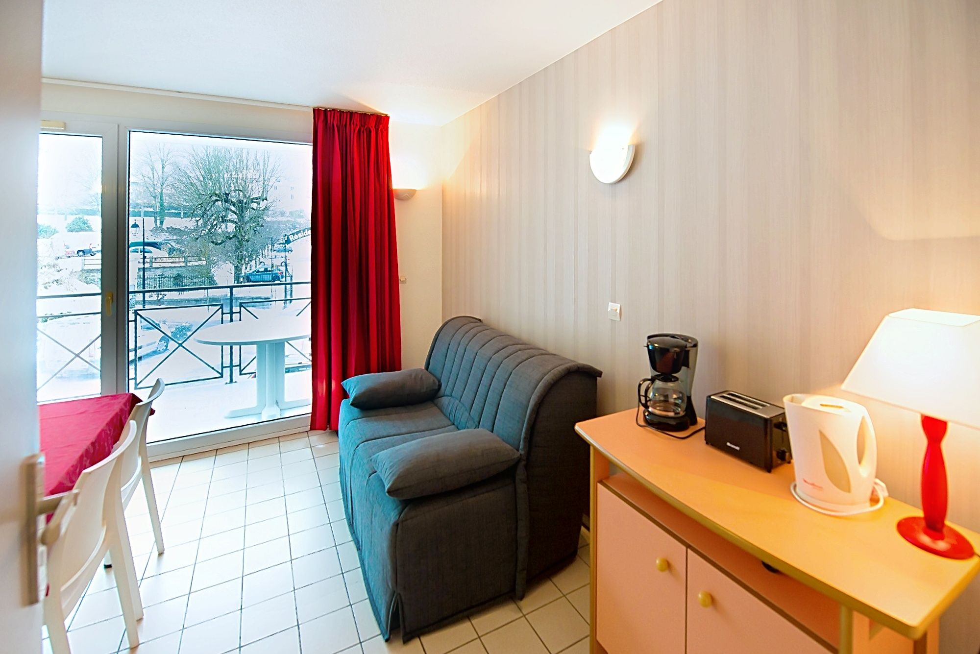 image Grey couch beside a large glass door with red curtains, bringing in the daylight.
