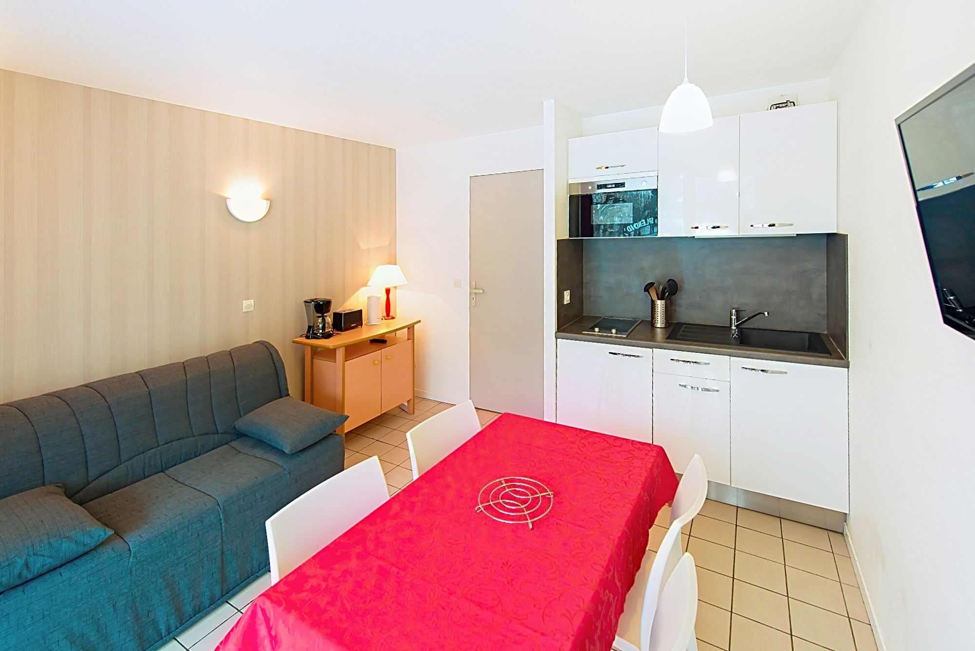 image Open living area with a grey couch, white kitchen and red tablecloth, made for sharing.