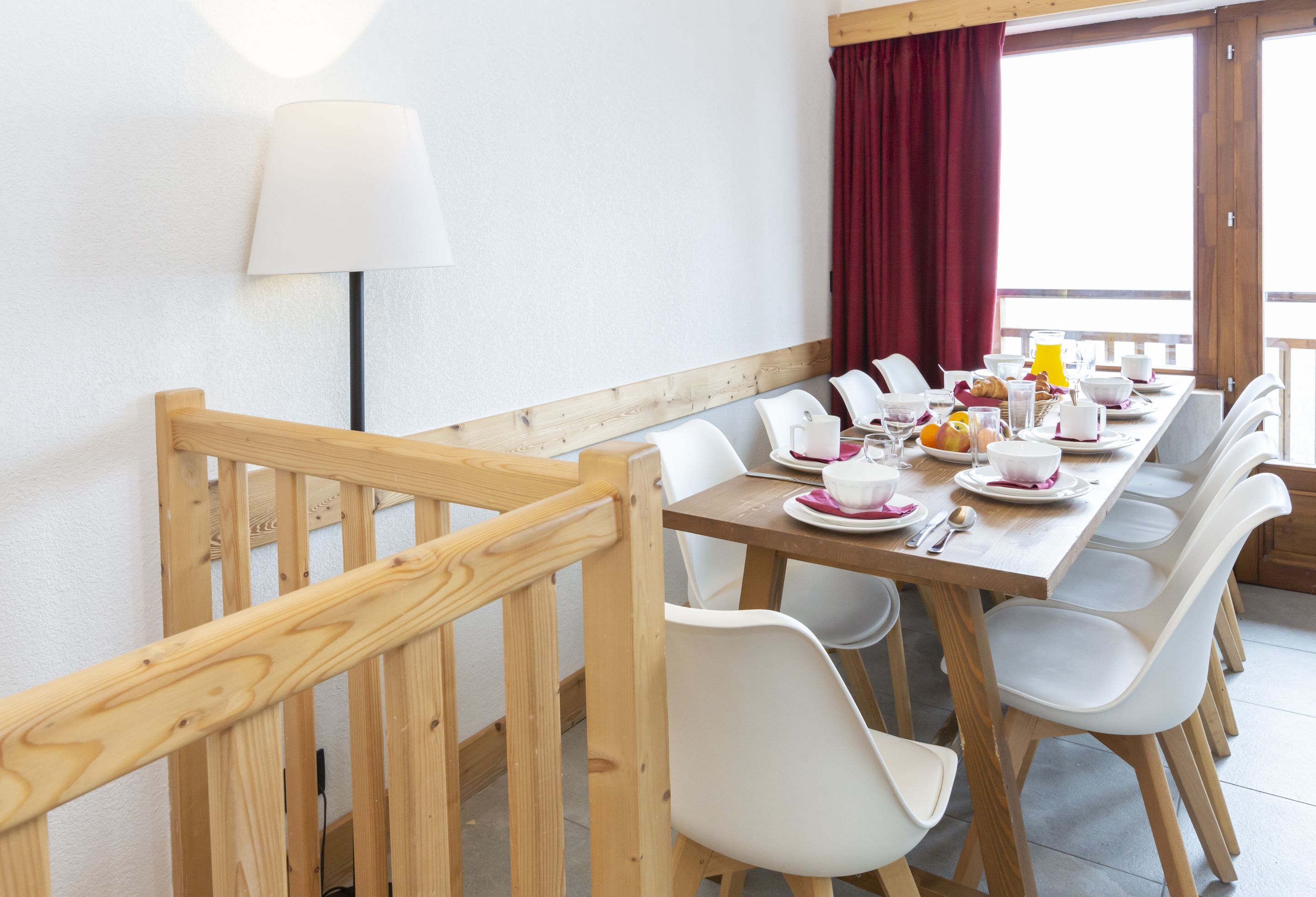 image The dining area set for ten, with natural light pouring through the red-curtained windows and the railing of the upper level visible beyond.