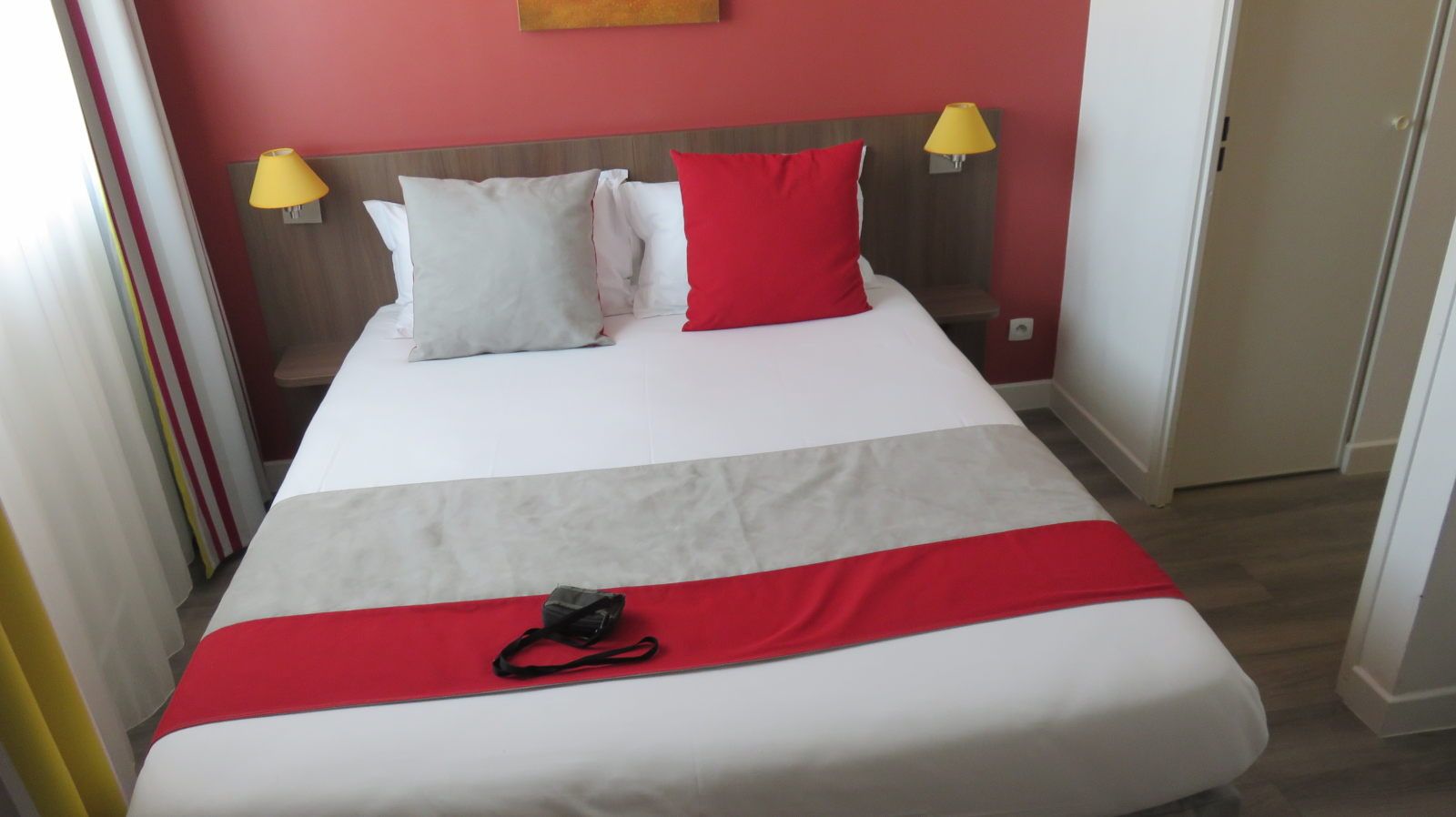 image Neatly made bed with red accent pillows against a wall and yellow bedside lamps