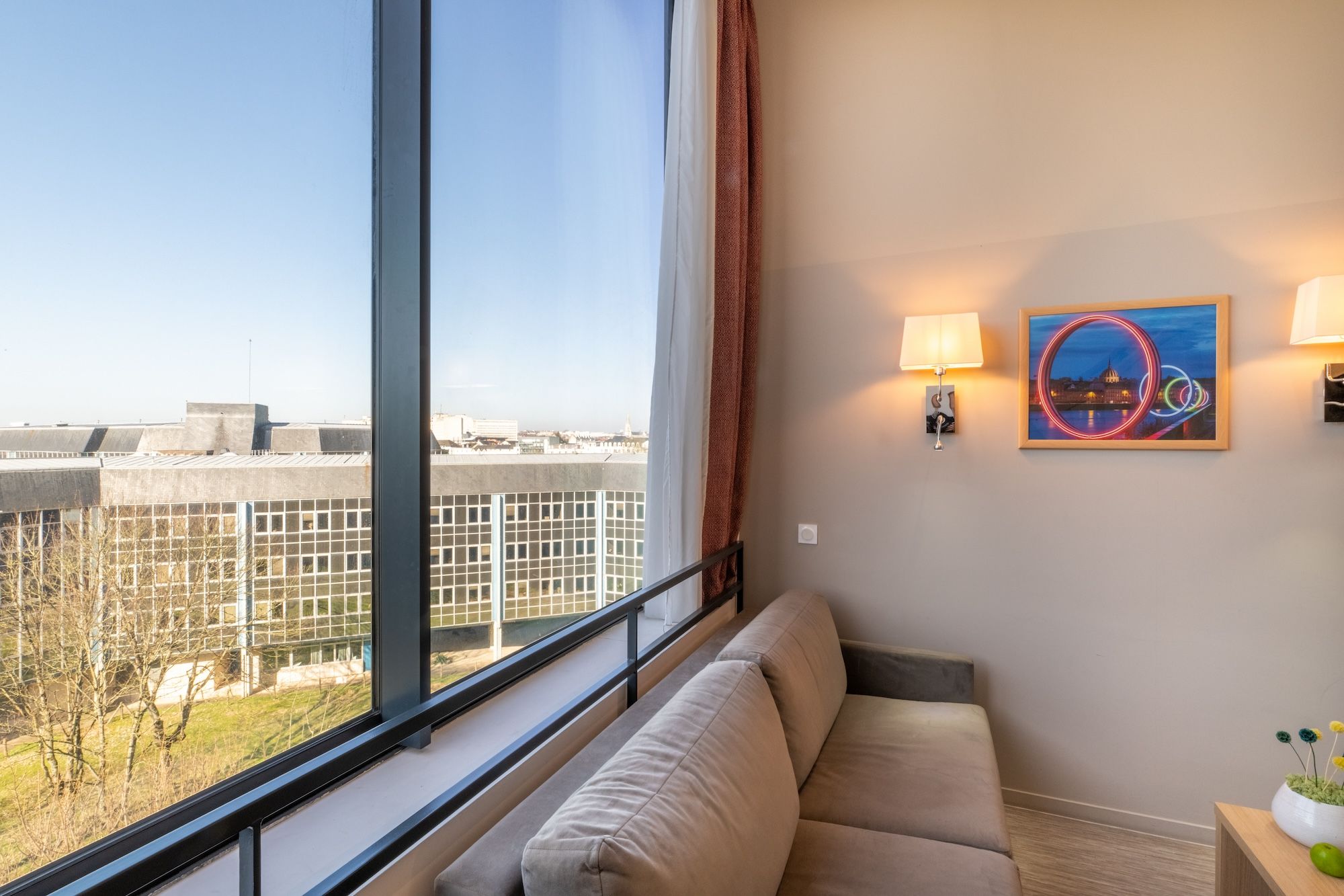 image A cozy living area featuring a soft gray sofa beside a large window, offering a bright view of the outside.