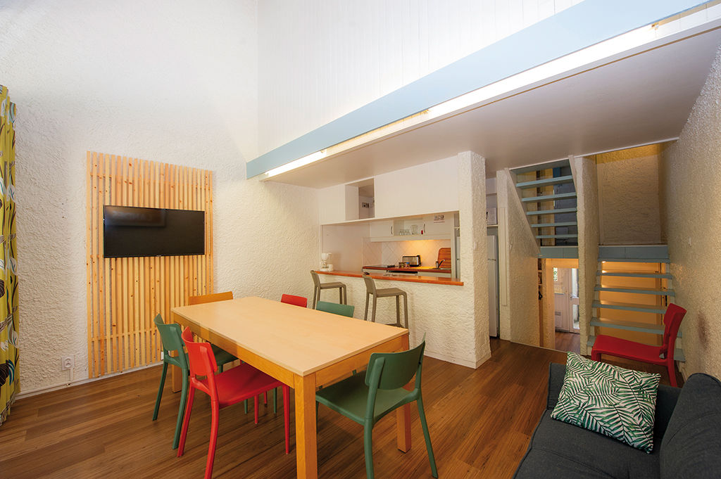 image Red and green chairs surround the wooden table, with a dark sofa, a wall-mounted TV and the kitchen and stairs behind