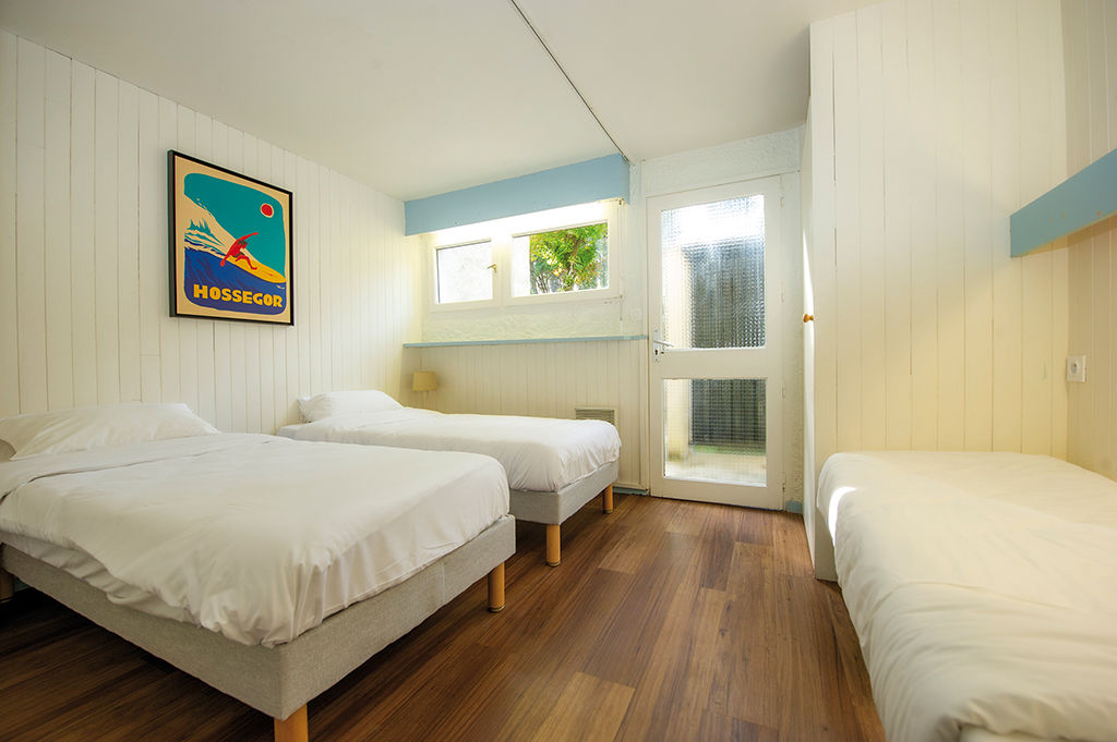 image Three single beds with white linens stand against white paneled walls, with a colorful poster and a window onto greenery