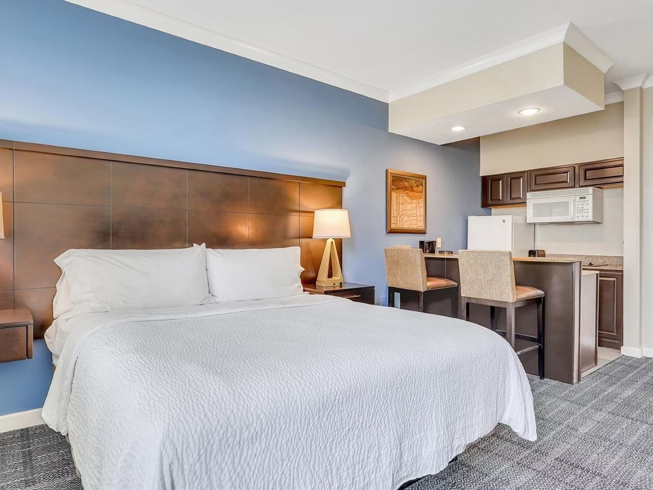 image Comfortable king bed with warm-toned linens, a lamp, and a built-in kitchen in view.