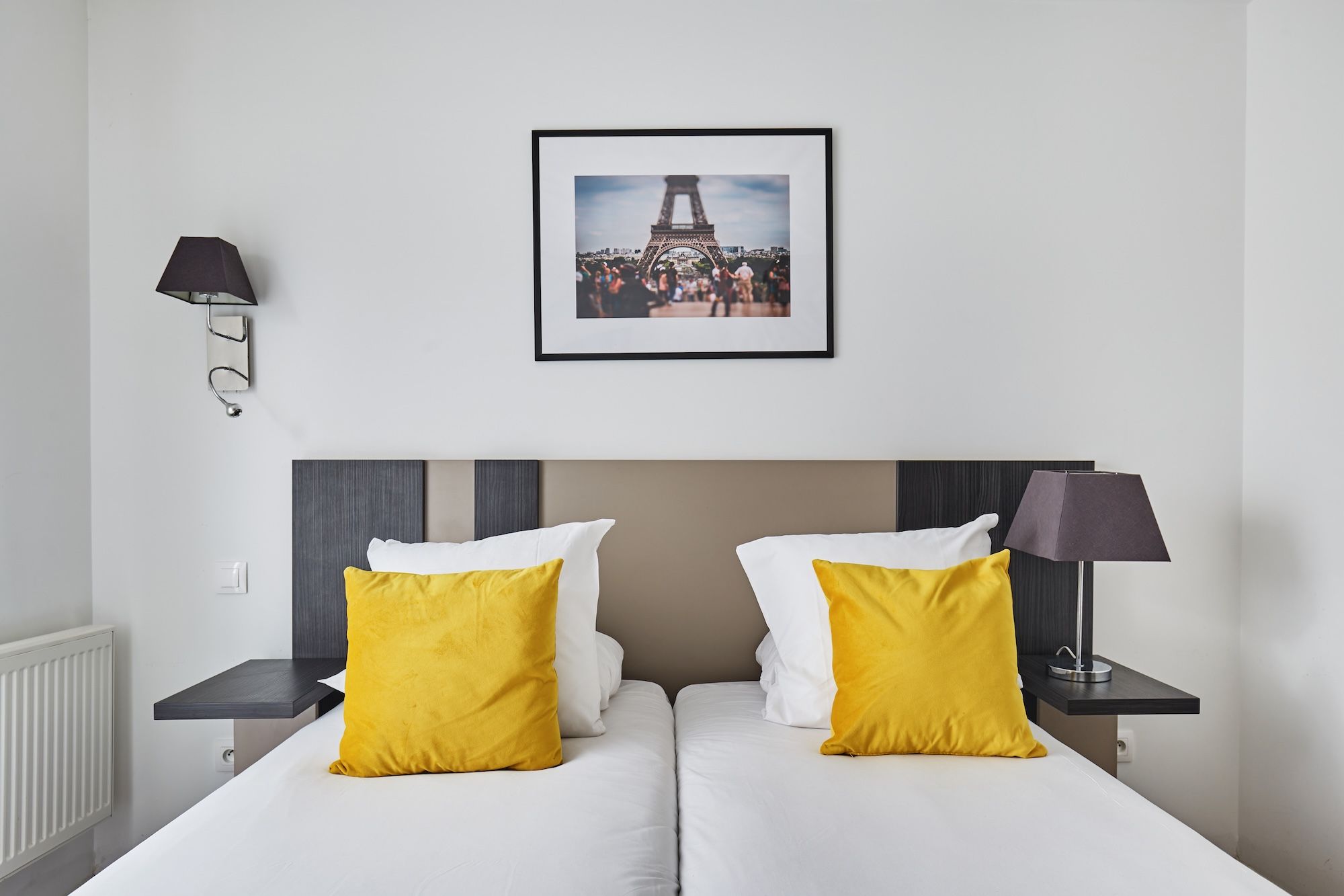 image This cozy studio features zip beds adorned with bright yellow pillows, complemented by modern wall sconces and a framed photograph of the Eiffel Tower.
