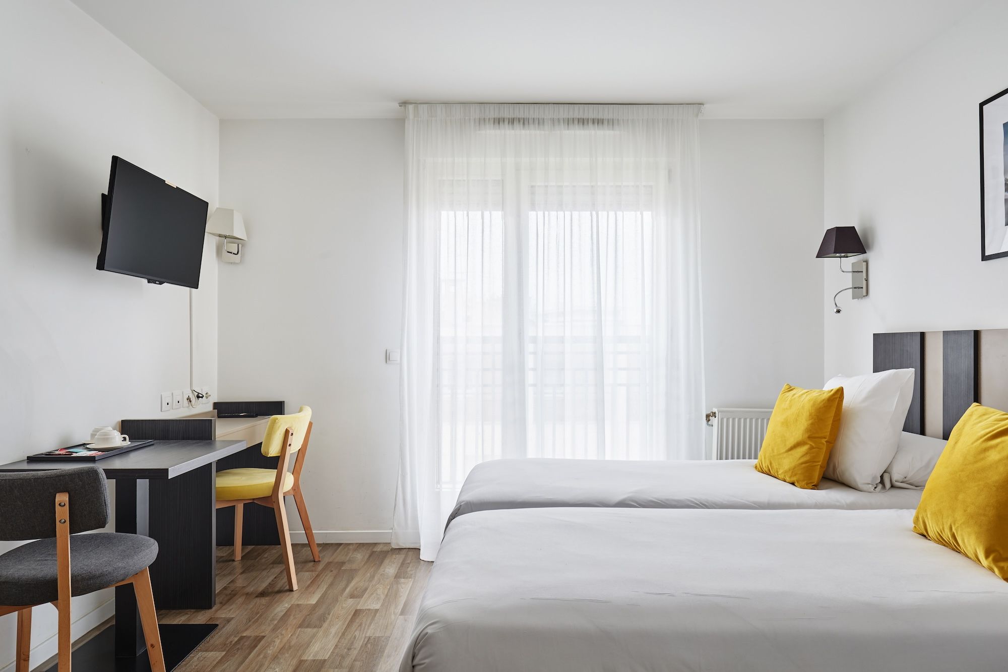 image Natural light filters through sheer curtains in this airy room featuring twin beds, a mounted TV, and a compact workspace perfect for any guest.