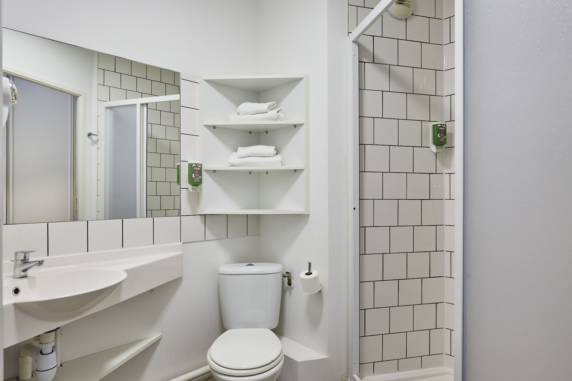 image This functional bathroom offers minimalist design with a pedestal sink, shower, and ample shelving for towels, all in a sleek tile design.
