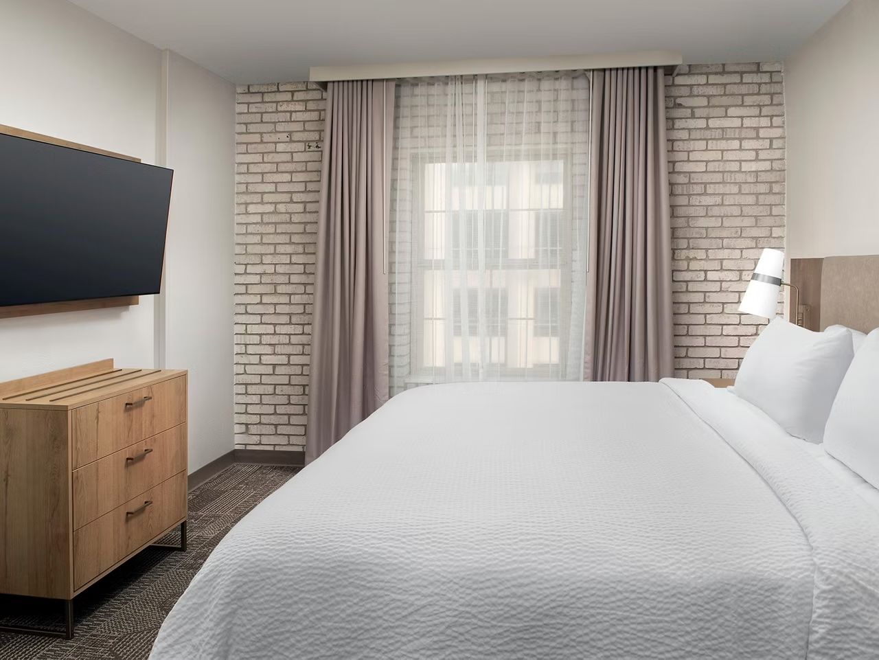 image King bed with white quilted bedspread, wall-mounted TV, wooden dresser, and a grey brick accent wall