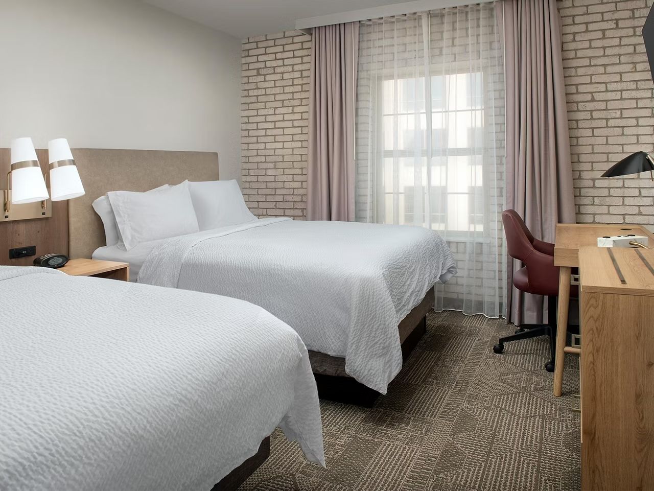 image Two queen beds with white bedding and light wood headboards, a wall-mounted TV, desk, and brick accent wall
