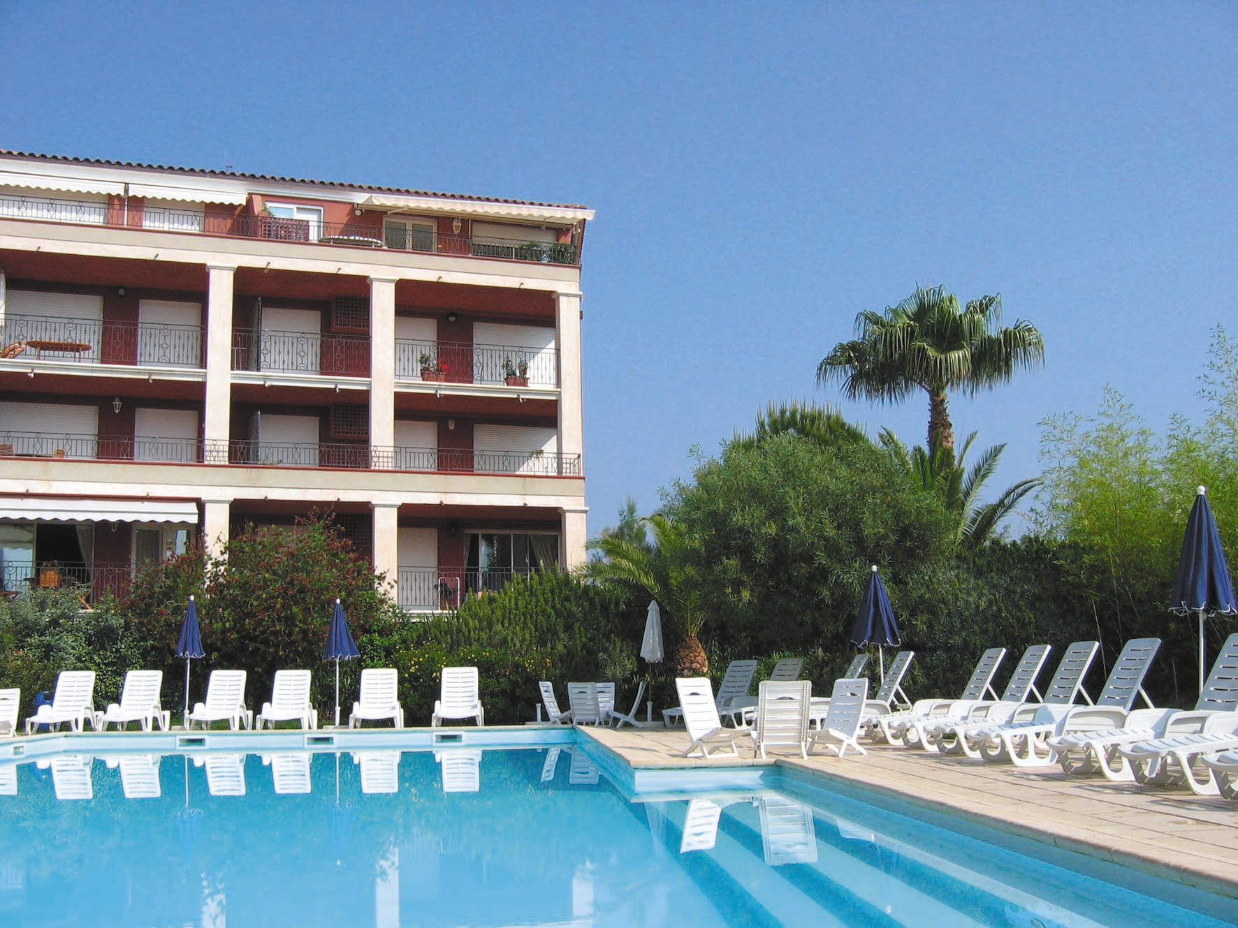 image nviting apartment complex with a large outdoor swimming pool, sun loungers, and palm trees under clear blue skies.