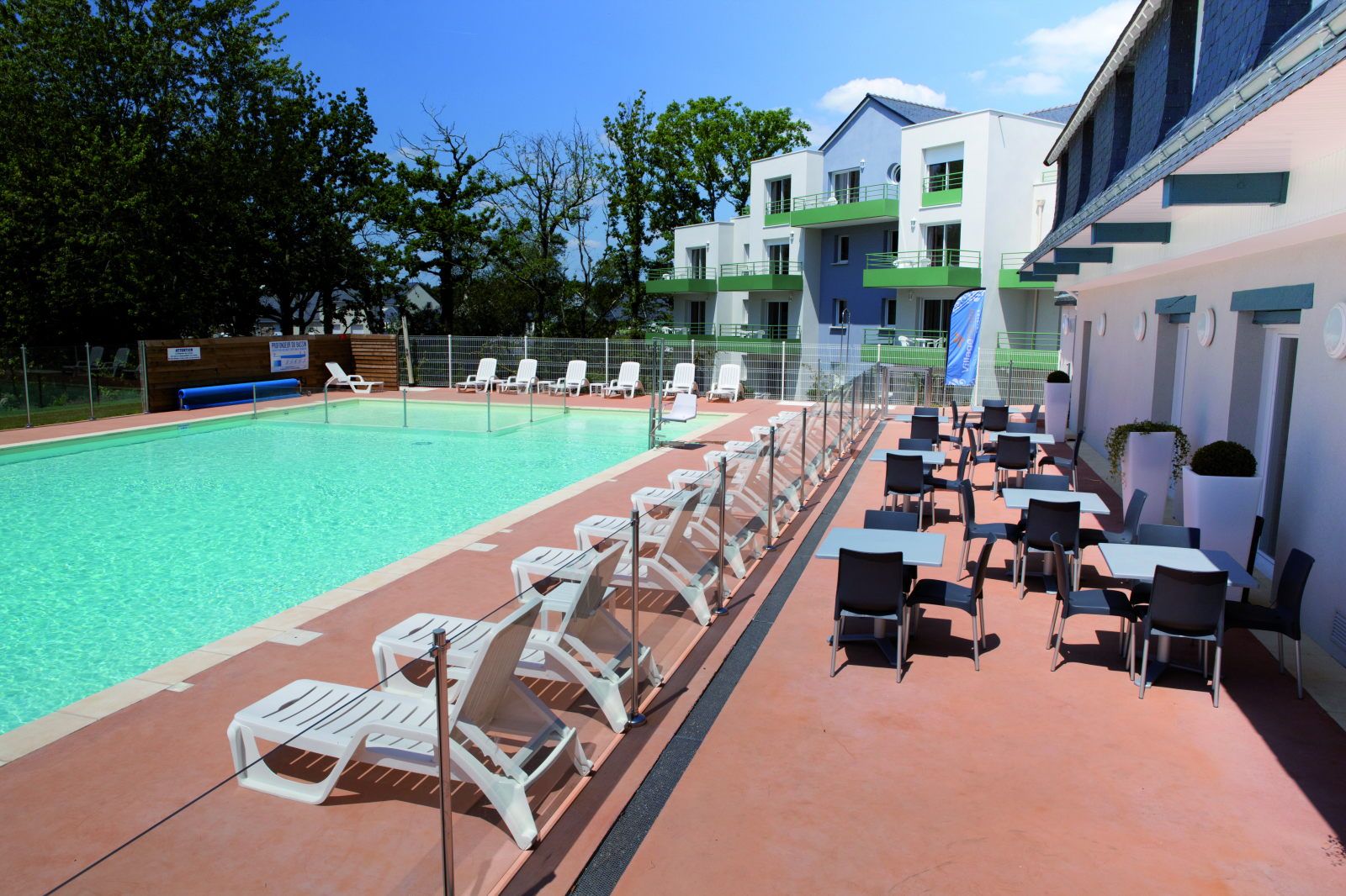 image Pool terrace with white loungers on one side and outdoor café tables and chairs on the other, apartment building behind