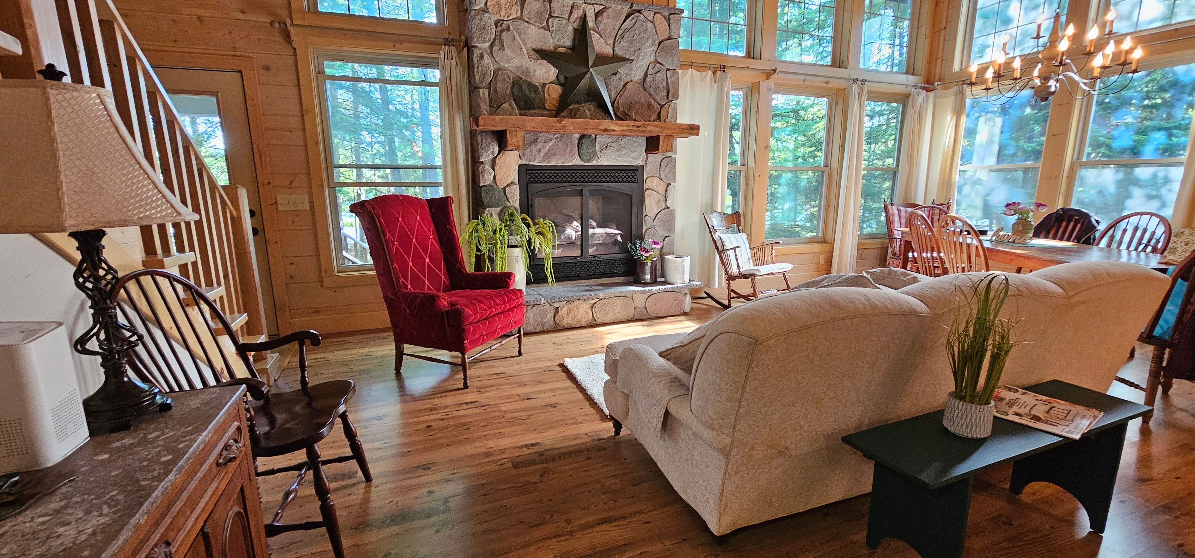 image Gather around the stone fireplace in this inviting living room with large windows offering stunning views of the surrounding nature.