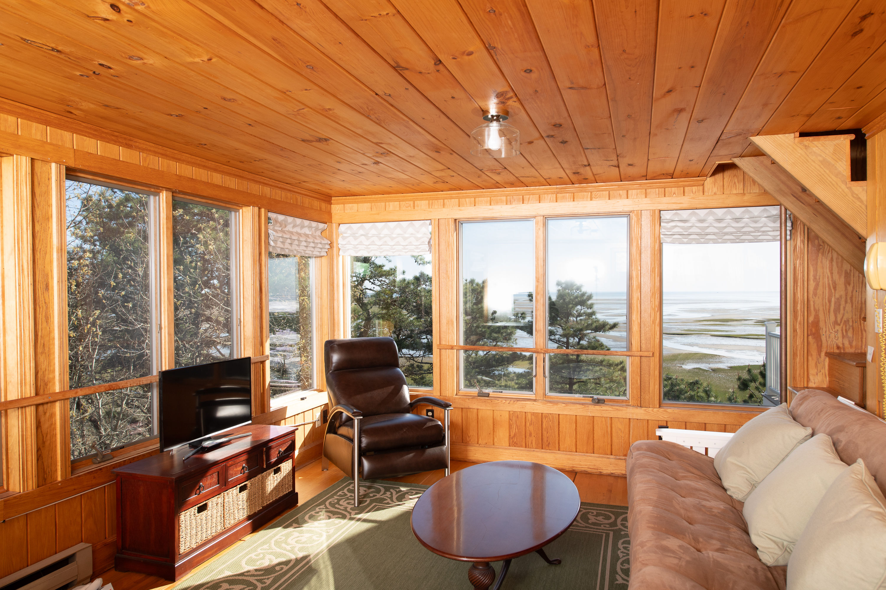image Versatile office/lounge space with wood paneling, featuring a TV, sofa, desk, and an elliptical bike, all while offering scenic water views.
