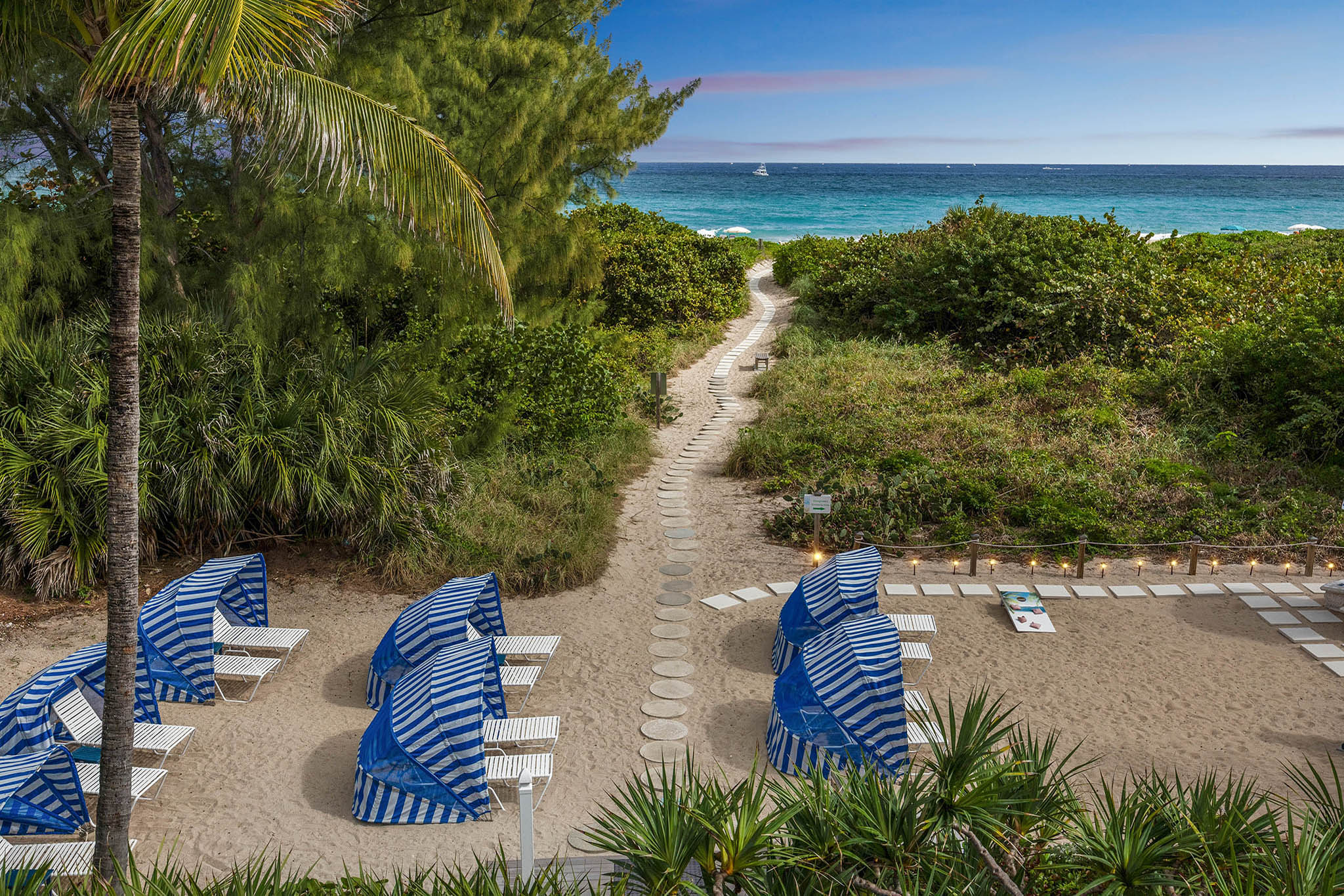 image A stone pathway bordered by palm trees and tropical plants leads toward the beach, with striped lounge canopies visible along the sandy shoreline at Sand Dune Shores.