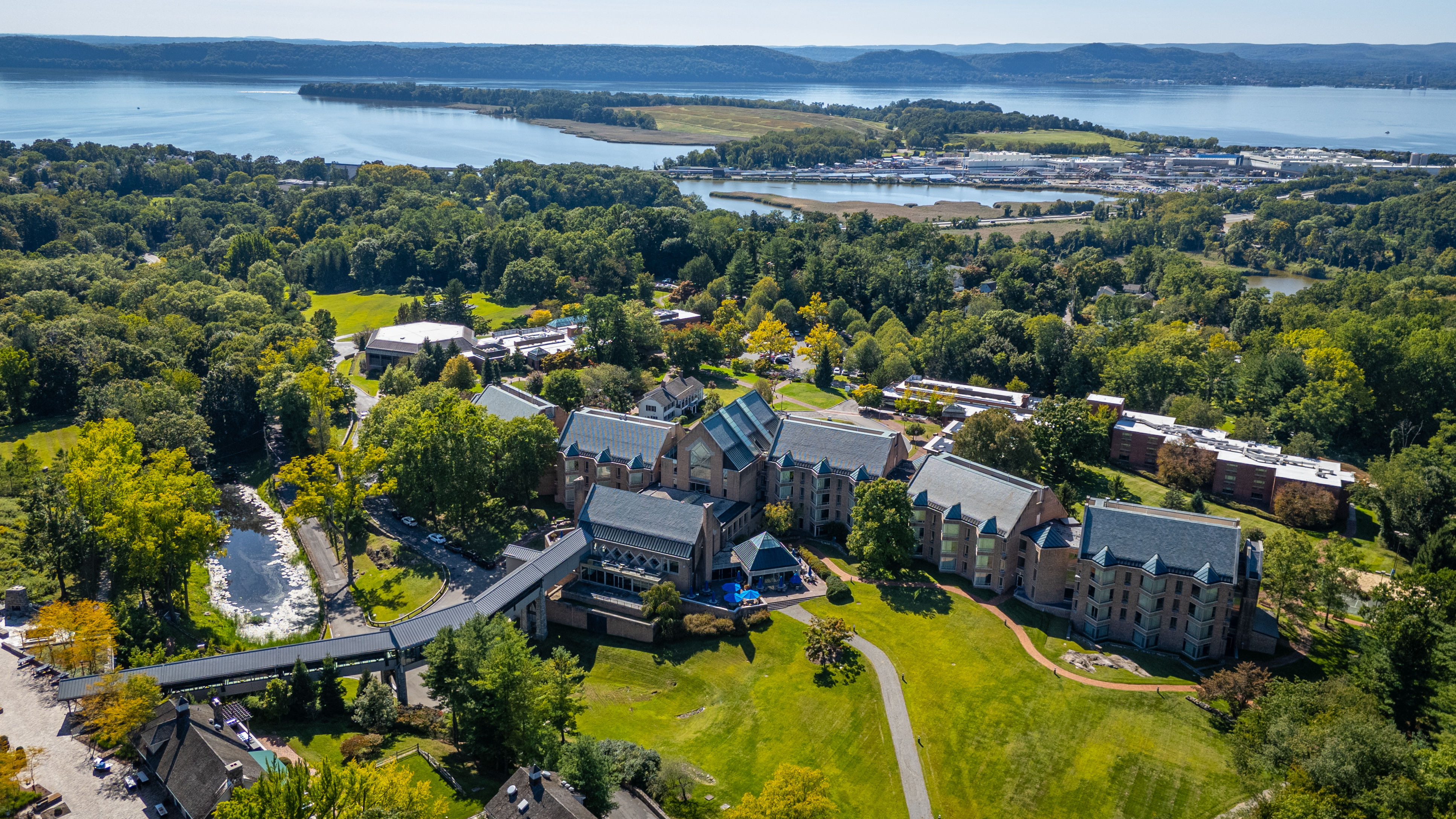 image A panoramic aerial view reveals the expansive grounds of Windrose on Hudson, nestled among lush greenery with the serene Hudson River creating a breathtaking backdrop.