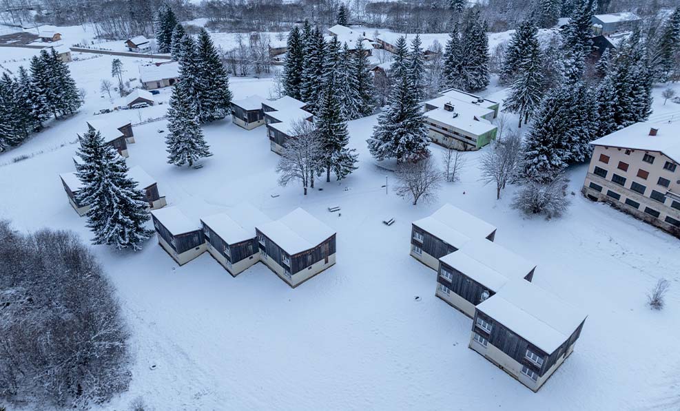 image Aerial view of Résidence Les Monts Jura, where cozy lodges are blanketed in soft snow amidst tall evergreen trees.