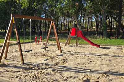 A sunny playground at Camping Verdon, featuring swings and a slide, surrounded by lush greenery.