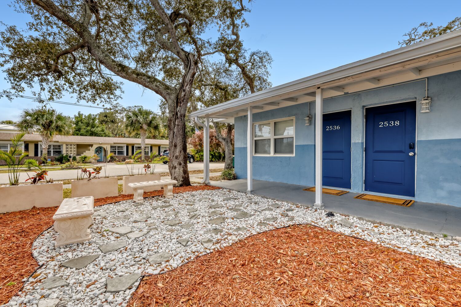 image Front entrance with two doors, a gravel walkway, and landscaped plants beneath shady trees.