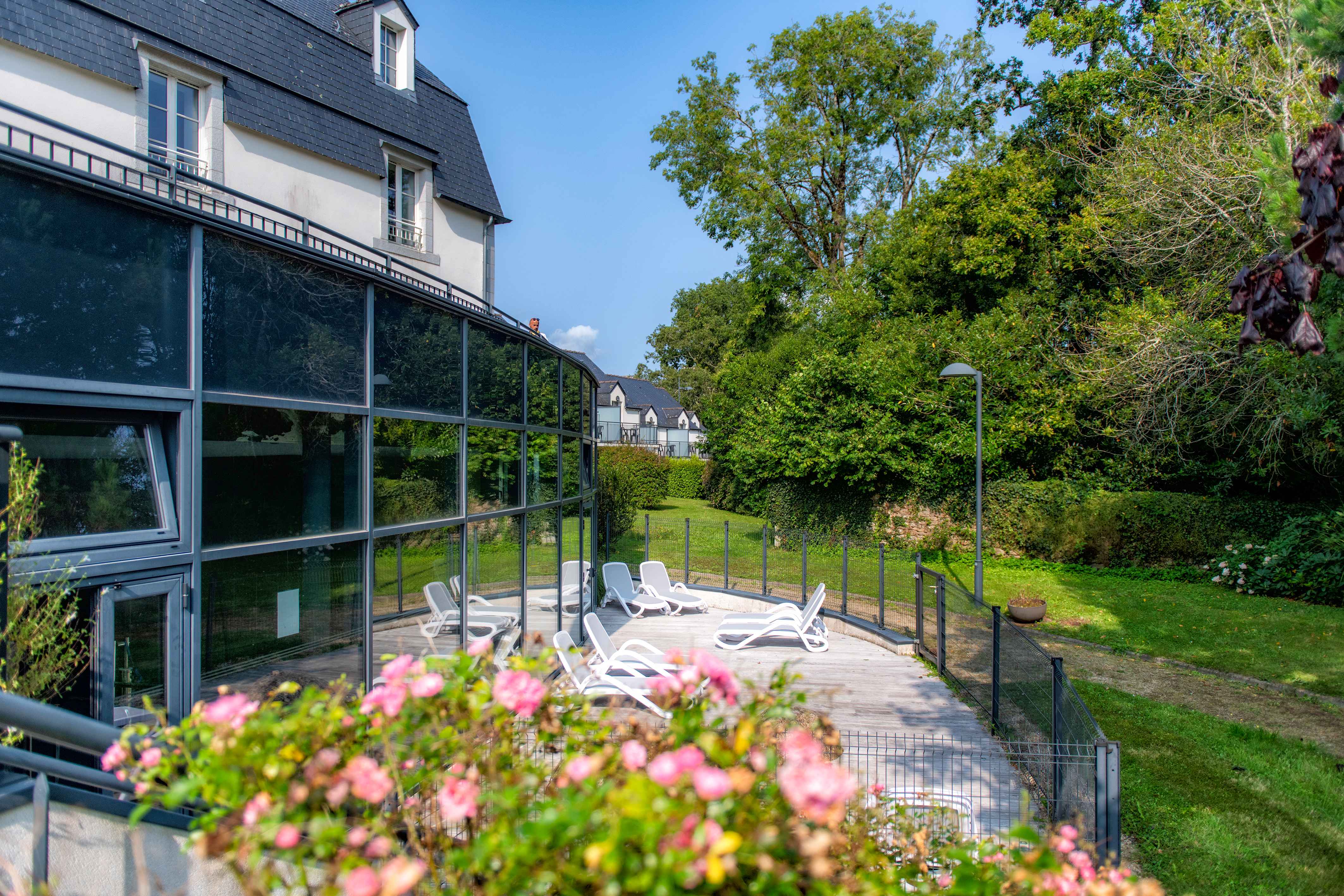 image White sun loungers line the poolside terrace, framed by trees and the glass walls of the pool building.