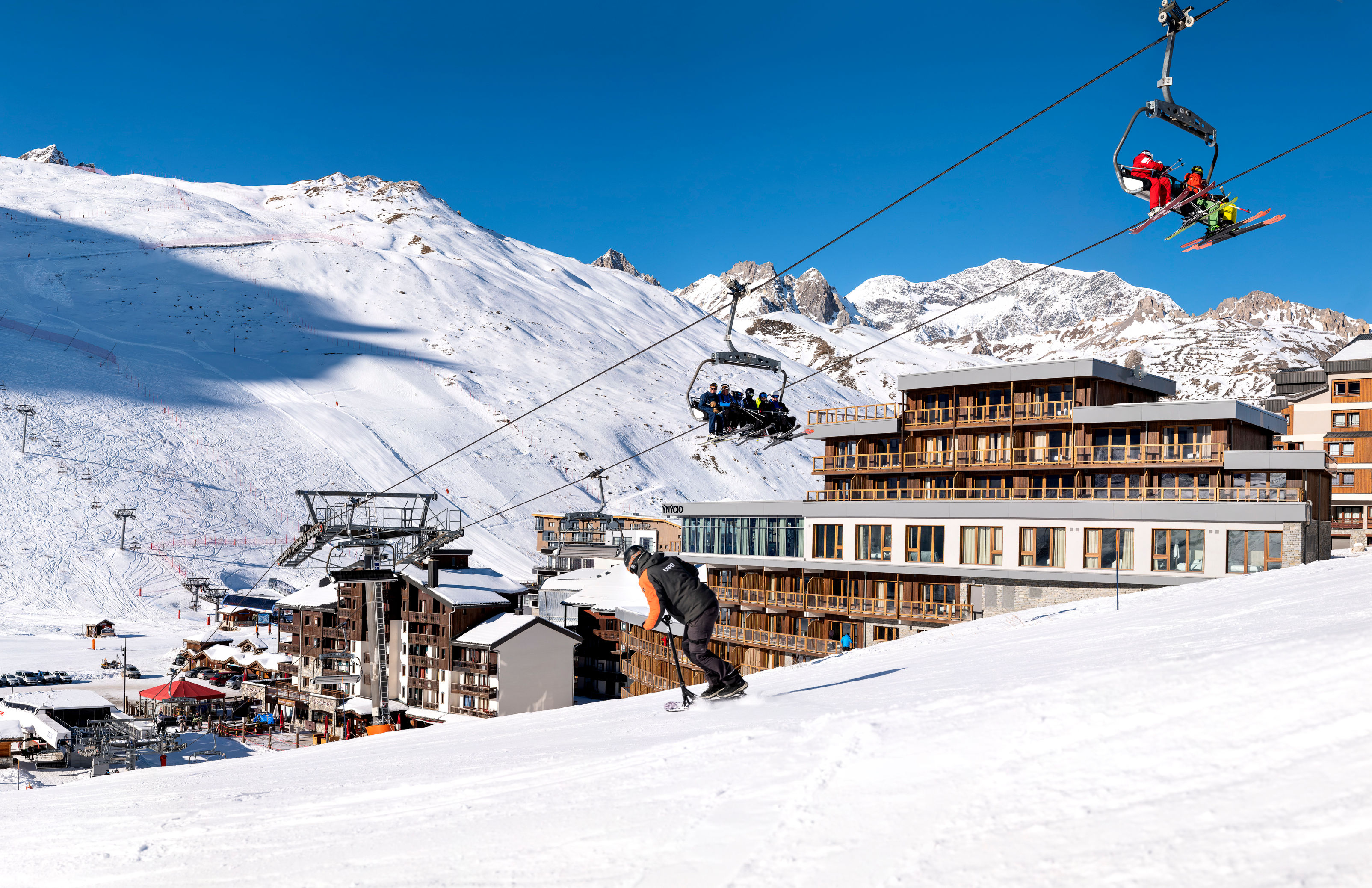 image A snowboarder carves down the piste below a chairlift, with village buildings and snowy peaks behind.