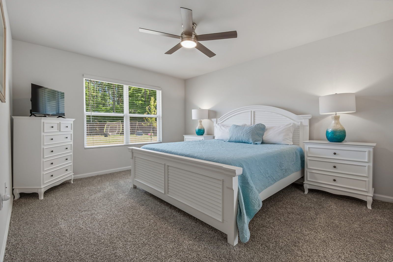 image White bed frame with light blue quilt, two nightstands with turquoise-base lamps, white dresser, and TV