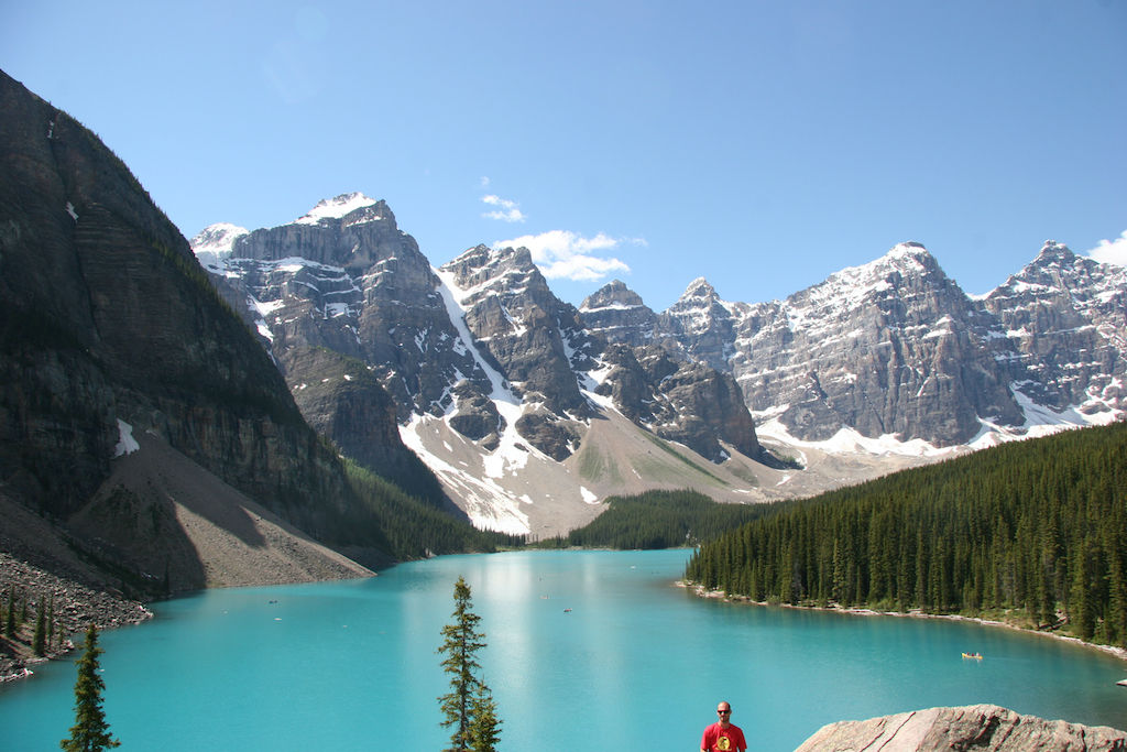 image Stand in awe at a breathtaking glacial lake, framed by snow-capped mountains and lush green pines under a clear blue sky