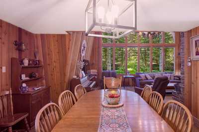 Long wooden dining table with eight chairs sits below a chandelier, forest visible through windows
