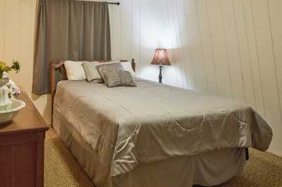 Gray quilt covers a wooden-framed bed beside a small side table and dark-shaded lamp