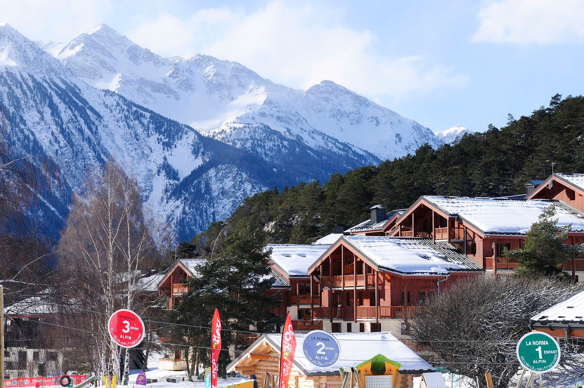 image Village of timber-clad buildings below rocky peaks, piste markers in front