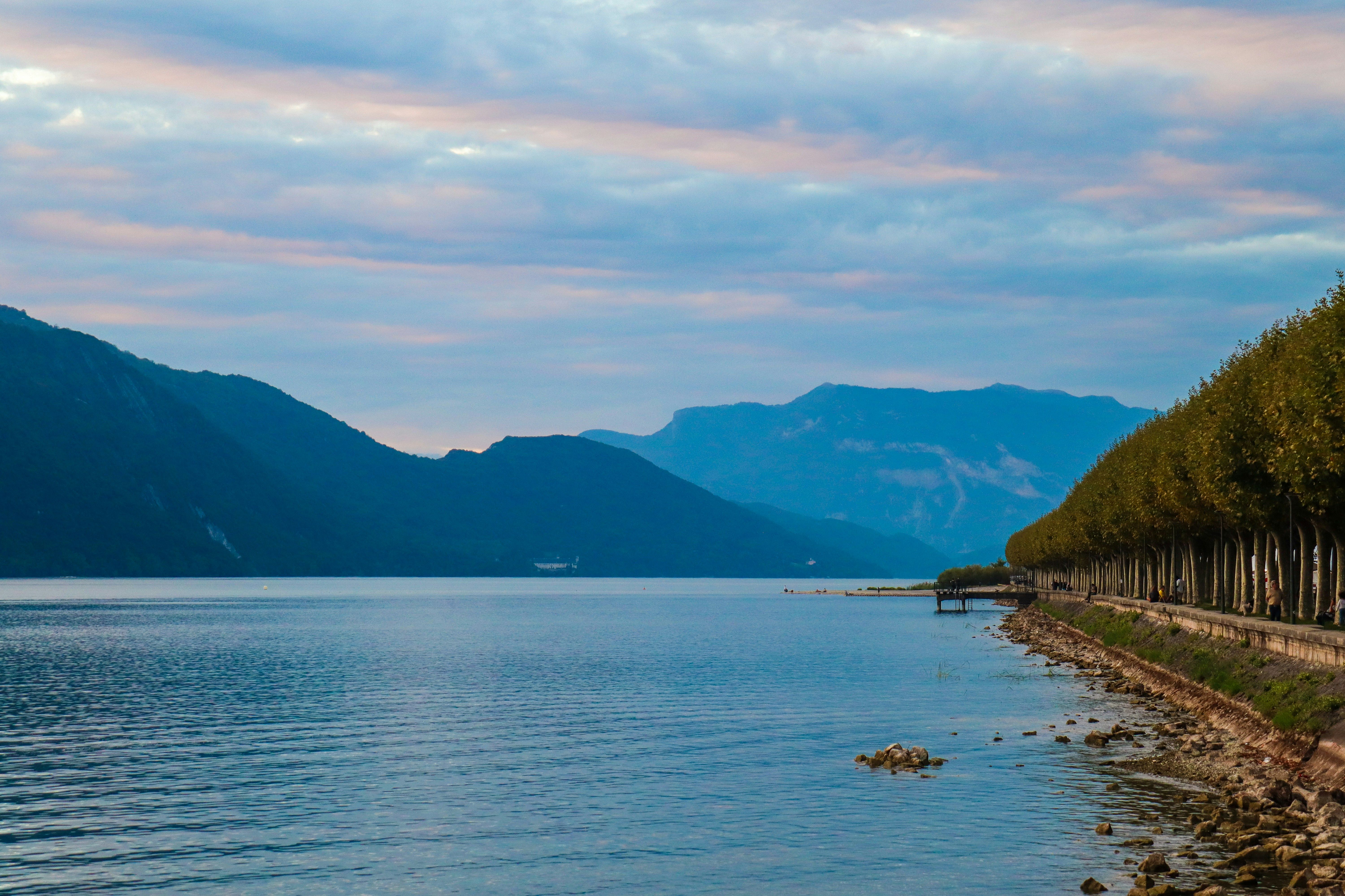 image Calm lake bordered by mountains and trees under a cloudy sky.