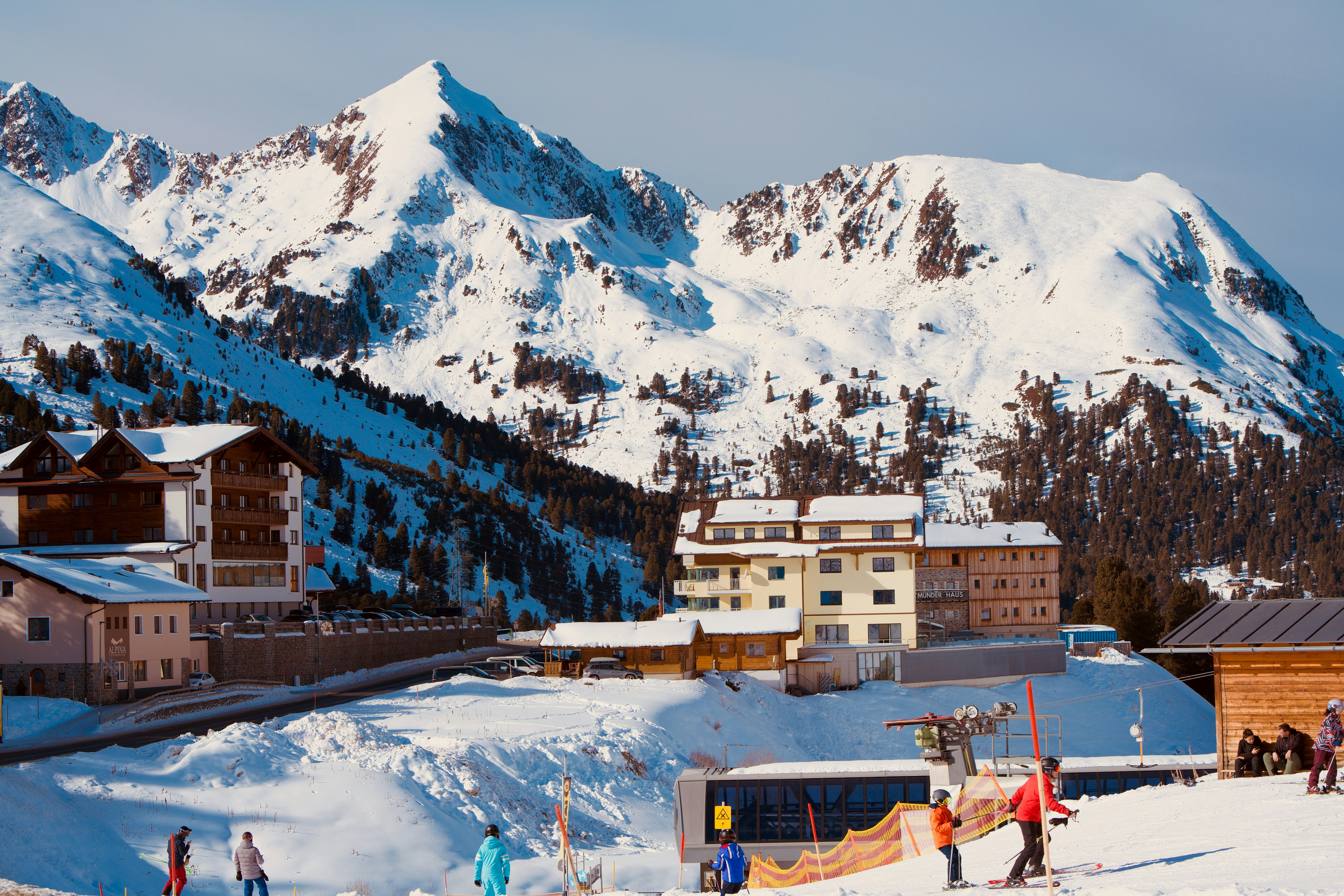 image Skiers glide across fresh snow beneath the lifts, with the buildings tucked against a dazzling mountain backdrop.